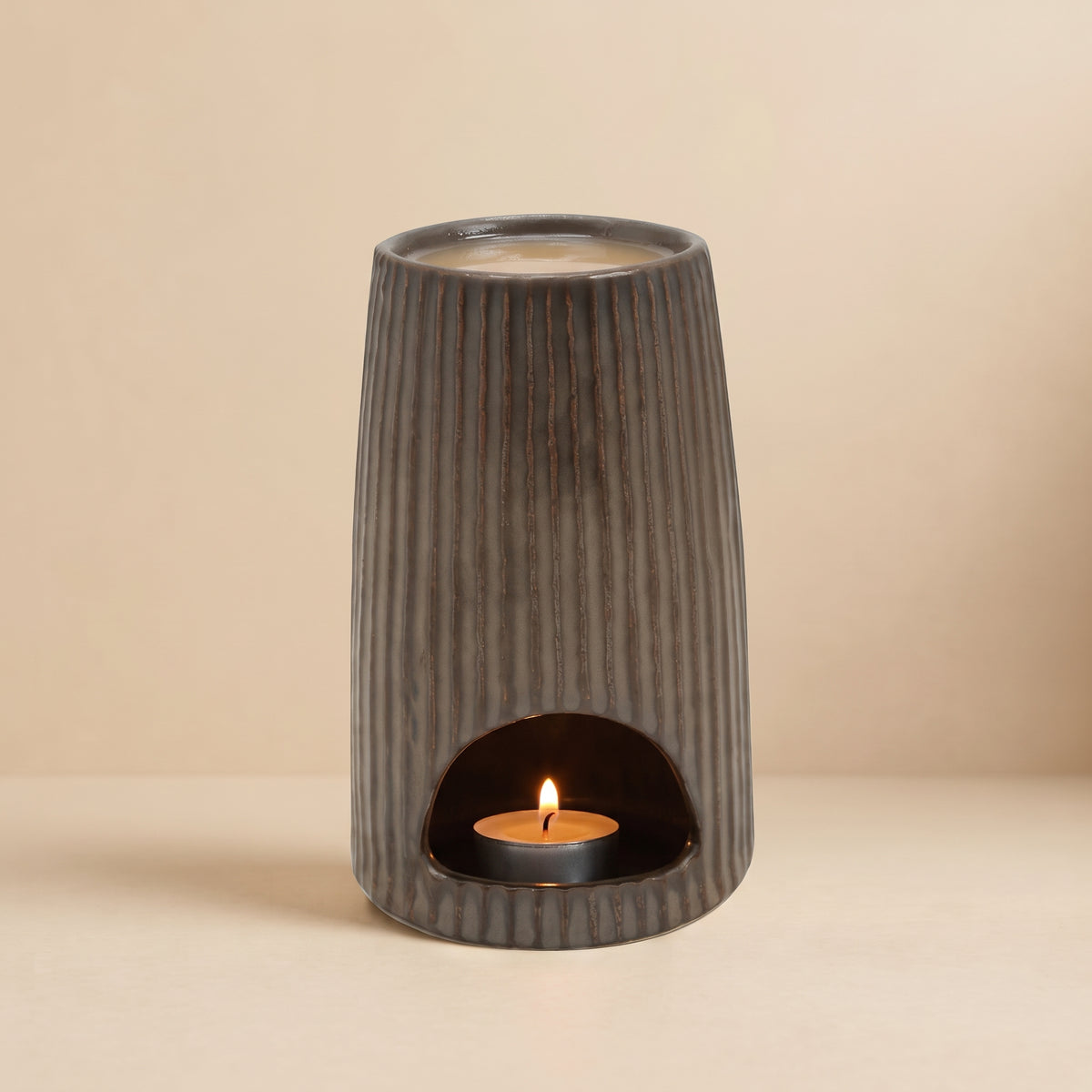 The glowing Bronze Ribbed Ceramic Wax Melt Burner by Hideaway Home Fragrances in a dark room, with a lit tealight inside and melting wax on top, diffusing cozy home fragrance.