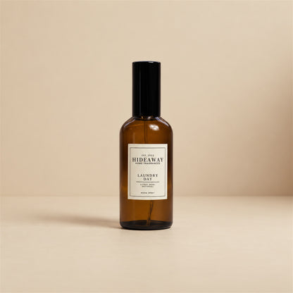 Hideaway Home Fragrances laundry day room spray styled on a wooden shelf next to folded white linen and fresh cotton towels.