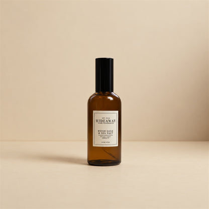 Hideaway Home Fragrances wood sage and sea salt room spray styled on a natural driftwood shelf beside white coastal pebbles and textured linen.