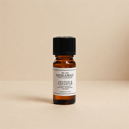Glass dropper dispensing Hideaway Home Fragrances orange and geranium fragrance oil into a ceramic wax burner beside fresh orange peel and botanical leaves.