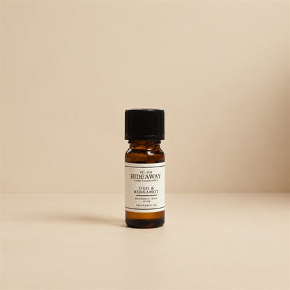 Glass dropper dispensing Hideaway Home Fragrances oud and bergamot fragrance oil into a ceramic burner beside cedarwood accents and warm spices.