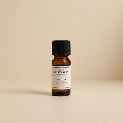 Glass dropper dispensing Hideaway Home Fragrances spa day fragrance oil into an aroma diffuser beside fresh eucalyptus sprigs and white rolled towels.