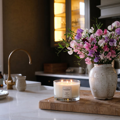 Hideaway Home Fragrances Sweet Pea 3 wick candle burning on a rustic wooden coffee table in a sunlit living room with floral decor