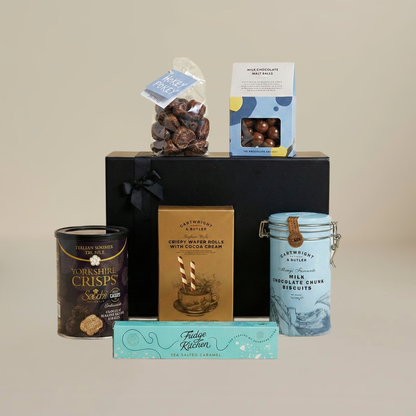 Luxury snack gift set flatlay with milk chocolate malt balls, wafer rolls, fudge slider, and buttery biscuits.