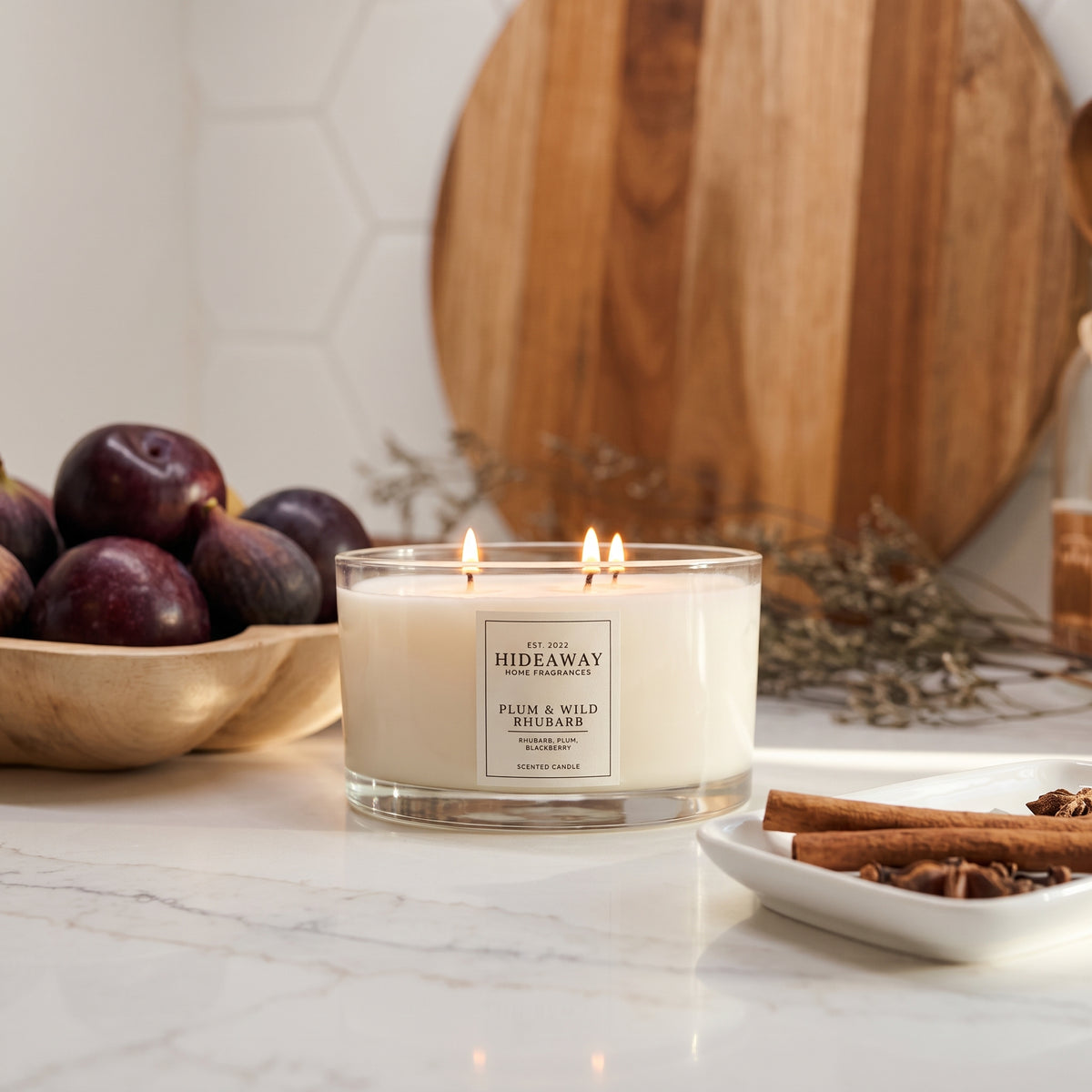 Hideaway Home Fragrances Plum and Wild Rhubarb 3 wick candle burning in a cosy living room setting on a rustic wooden coffee table surrounded by home decor